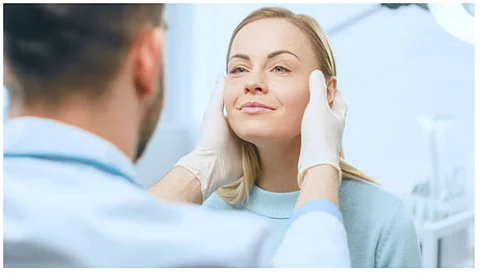 A woman with blonde hair is getting her face examined by a professional wearing glove.