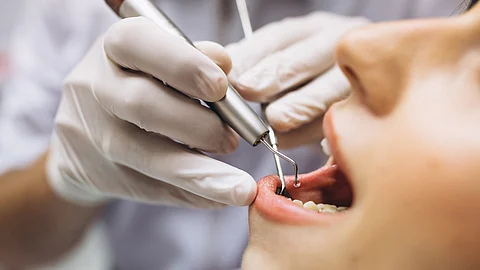 A woman sits in a dental chair while a dentist cleans her teeth with dental tools.