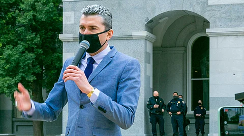 A man in a blue suit speaks into a microphone while police officers stand watch in the background outside a building.