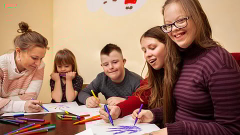 Image shows 3 children and 2 adults in a daycare facility painting with color pencils.