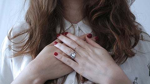 A woman in a white shirt with red nails holds her hands to her chest, expressing concern about stress cardiomyopathy