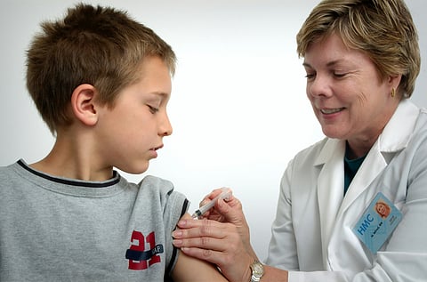 A nurse in a white coat administers a vaccine to a young boy wearing a grey T-shirt.