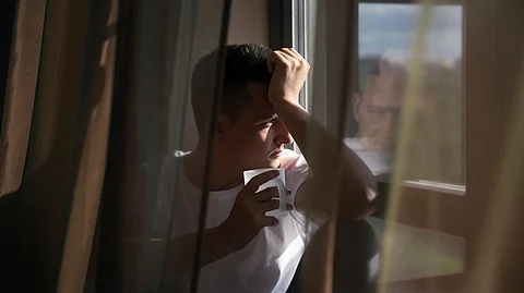 A man in a white t-shirt holding a cup, sitting by a window with his head resting on his hand.