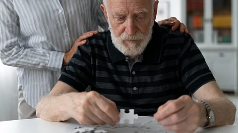 An elderly man trying to do a jigsaw puzzle with a bystander nearby.
