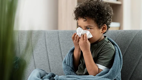 A curly haired boy blowing his nose using a tissue while sitting on a sofa covered with a blanket.