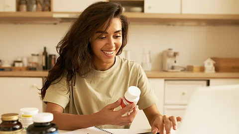 Young female holding skin supplement in one hand and researching about it on laptop.