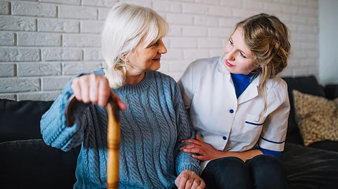 An elderly lady talking to a nurse sitting on a couch clutching a walking stick.