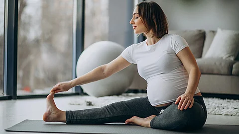 A pregnant woman stretching out her legs and arms in a white t-shirt and grey joggers.