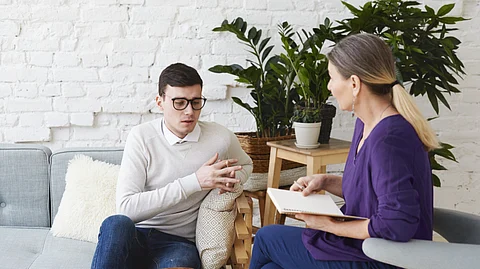 Image shows a man and woman seated on different couch, engaged in conversation about reshaping addiction treatment.