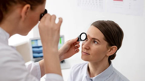 Image shows an eye doctor examining a patient's eye using magnifying lens in a clinical setting.