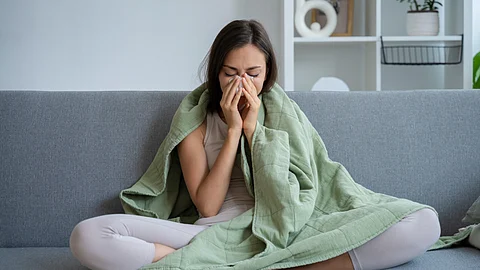 A woman sitting on a couch, covered with a blanket, pinching her nose as she is sick.