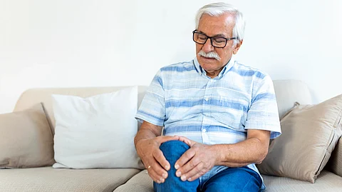 Image shows elderly man sitting on a couch, holding his knee in discomfort.