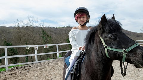 Young girl on a black pony with a wheelchair beside her, outdoors under a blue sky.