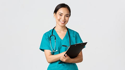 A healthcare professional in blue medical scrubs, wearing a stethoscope and holding a clipboard.