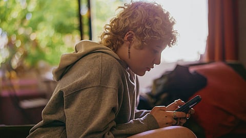 Image of a young boy having light brown curly hairs using a mobile phone while sitting.