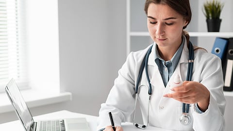 Doctor writing a medical prescription at desk with laptop and medicine bottle.