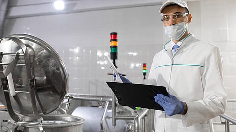 Quality control inspector wearing protective clothing, mask, gloves, and safety glasses, holding a clipboard while examining equipment in a processing plant.