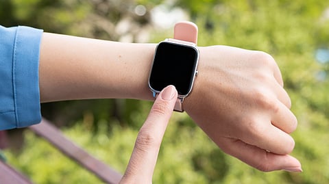 A person wearing a blue sleeve and a pink smartwatch with a black screen on their wrist, touching the screen with their finger, with a blurred green outdoor background.