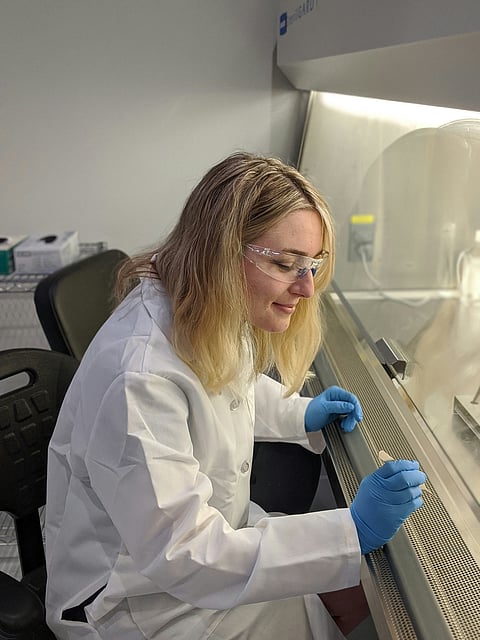 A woman in a lab coat examines a piece of glass, focused on her work in a laboratory setting.