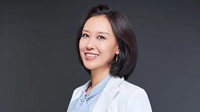 A woman wearing a white blazer over a black dress, standing confidently with a neutral background.