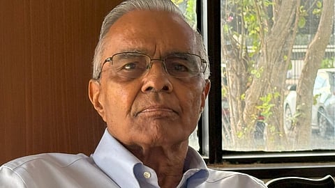 Image of an elderly man with gray hair wearing a light blue button-up shirt, seated indoors near a window.
