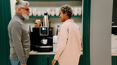 A man and woman stand together in front of a coffee maker, smiling as they prepare to brew coffee.