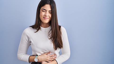 A young brunette woman stands against a blue background, holding her stomach in pain from IBS.