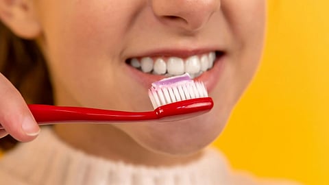 A smiling person holding a red toothbrush with toothpaste, illustrating the debate on brushing before or after breakfast for better enamel protection.