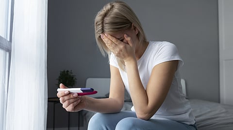 Image of a woman sitting on a bed, holding a pregnancy test in one hand, covers her face with the other.