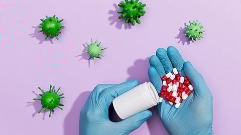 Image of a capsules of red and white color in hands of a person wearing surgical gloves along with graphics of bacteria.