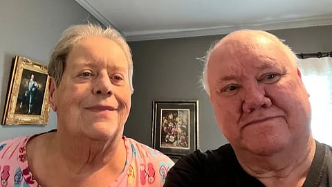 Elderly couple smiling indoors, with gray walls and framed paintings in the background.