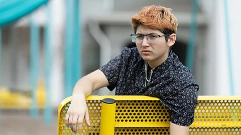 Image of a teen with short peach orange hair and glasses leaning on a yellow perforated metal bench.