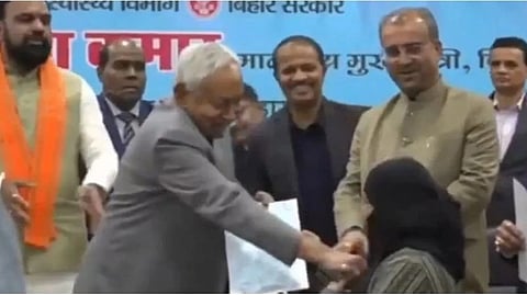 An image of a group of people stand on a stage, smiling. An older man in a gray suit hands a certificate to a seated woman in black.