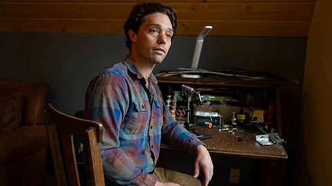 An image of a man in a plaid shirt sits at a cluttered wooden desk with tools.