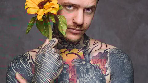 An image of tattooed man holds a sunflower against a gray backdrop.