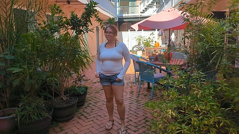 An image of a woman stands in a courtyard with brick flooring, surrounded by lush potted plants.