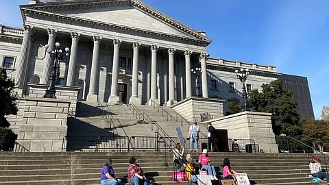 People gather on the steps of a grand building with columns, holding signs.