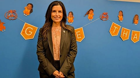 A woman in a suit smiles in front of a blue wall decorated with a festive banner and caricatures.