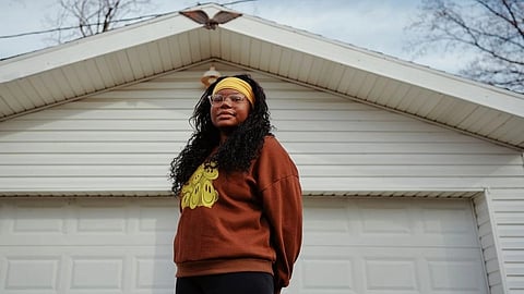 A woman in a brown sweater with yellow patterns and a yellow headband stands in front of a white garage door.