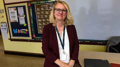 A woman with glasses and a badge, wearing a maroon blazer, sits in a classroom.