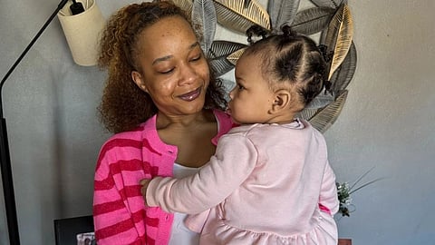 A woman in a pink striped sweater lovingly holds a toddler in a pale pink outfit.