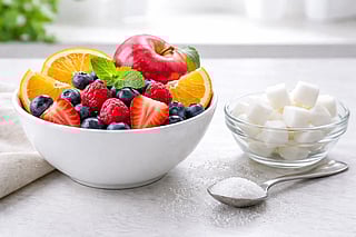 A large bowl containing a variety of fruits and a smaller bowl with sugar cubes, with a spoon filled with sugar placed beside it
