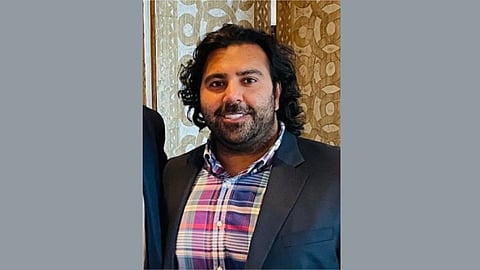 Portrait of Anosh Ahmed wearing a checkered shirt and a blue blazer, smiling while posing.