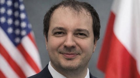 Close-up of a smiling man in formal attire with a blurred background featuring two national flags.