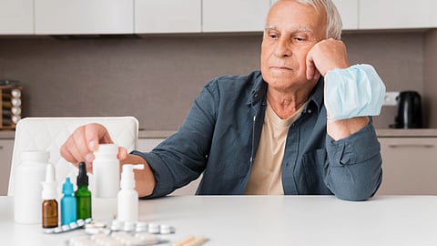 An elderly man sits at a kitchen table, thoughtfully looking at several medication bottles and pills.