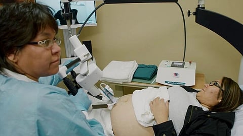 A healthcare professional in blue scrubs prepares an ultrasound machine beside a pregnant woman.