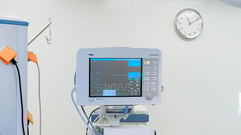 A hospital room with a monitor showing vital signs, connected cables, and a wall clock set at 10:10.