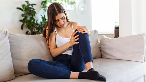A woman sitting on a sofa wearing a white tank top and jeans, holding her knee in discomfort.