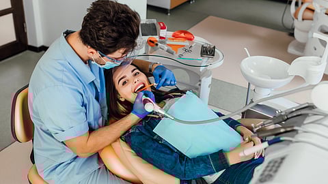 A dentist in blue scrubs, wearing gloves and a mask, cleans a patient's teeth in a clinic.