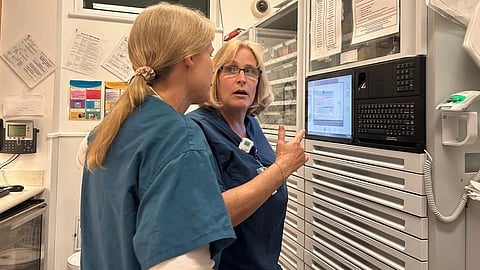 Two medical professionals in scrubs discuss something at a computer terminal in a hospital setting.
